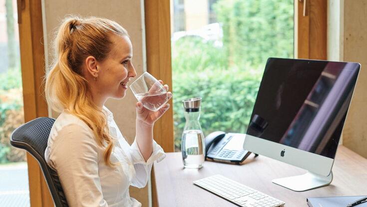 Frau sitzt am Schreibtisch mit einem Glas Wasser in der Hand