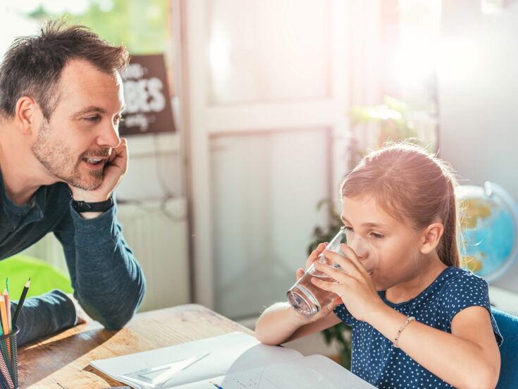 Ein kleines Mädchen trinkt mit beiden Händen ein Glas Wasser und ihr Vater schaut ihr dabei zu.