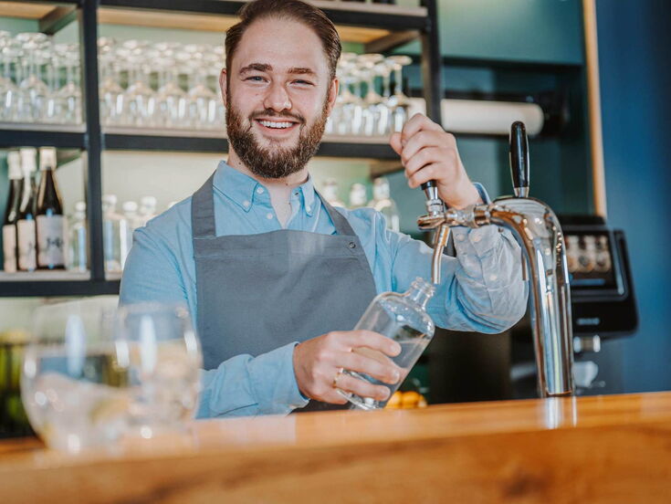 Ein Mann steht hinter einer Theke uns zapft Wasser in ein Glas aus einer Zapfanlage. Er lächelt dabei in die Kamera.