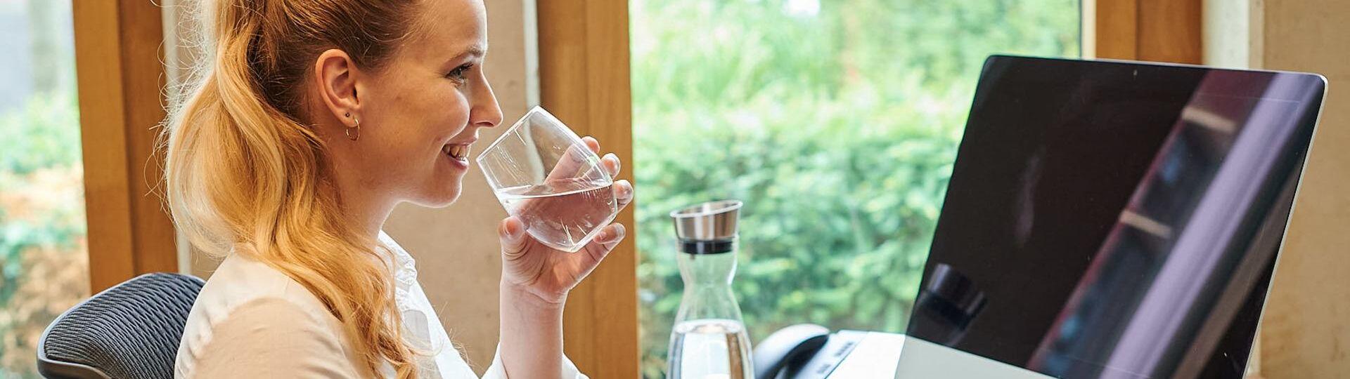 Frau sitzt am Schreibtisch mit einem Glas Wasser in der Hand